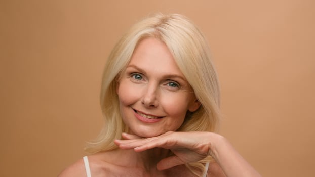 Close up portrait in studio beautiful mature old woman Caucasian model touching soft moisturized skin face smiling advertise of anti-aging lifting beauty cosmetics wrinkle skincare beige background photo