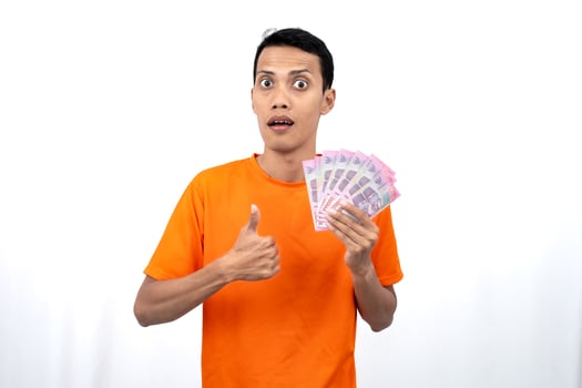 Portrait of a young Asian man wearing an orange t-shirt with a happy, surprised, pleased expression while carrying hundreds of thousands of rupiah. isolated on a white background. photo