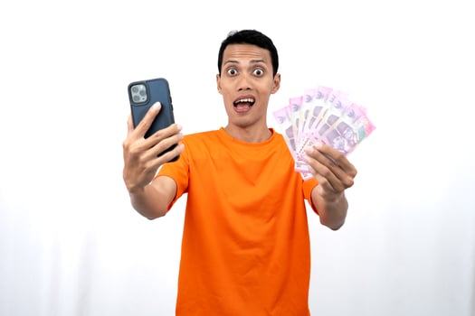 Portrait of a young Asian man wearing an orange t-shirt with a happy, surprised, pleased expression while carrying a smartphone and hundreds of thousands of rupiah. isolated on a white background. photo