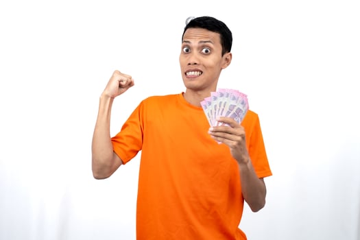 Portrait of a young Asian man wearing an orange t-shirt with a happy, surprised, pleased expression while carrying hundreds of thousands of rupiah. isolated on a white background. photo