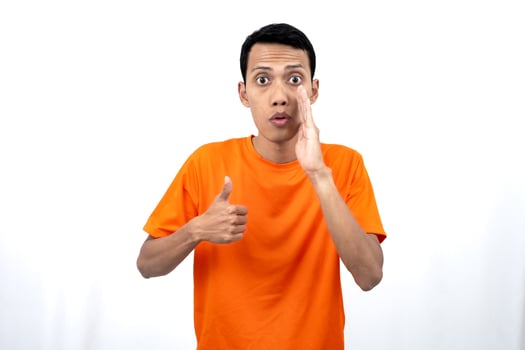 Portrait of a young Asian man wearing an orange t-shirt with his hand near his mouth whispering gossip and telling secrets while sharing news isolated on white background. photo