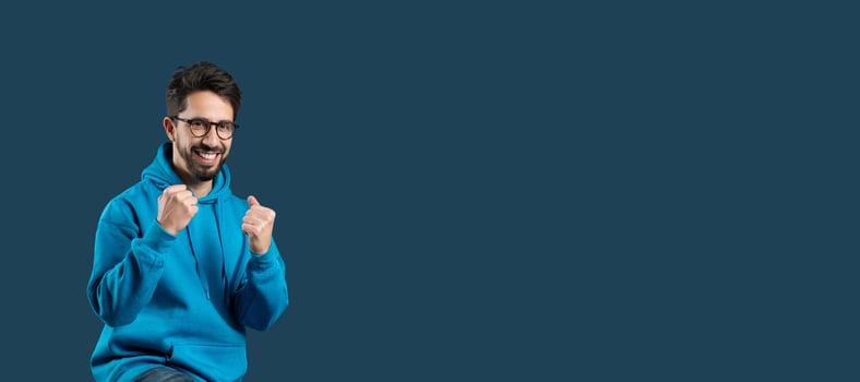 A young man with beard and wearing glasses clenching his fists in a gesture of victory and excitement. He is dressed in a casual blue hoodie and stands against an isolated blue background, copy space photo