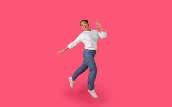 Asian young woman jumps and smiles in front of a bright pink background, her arms outstretched and her white shirt billowing in the air. photo