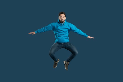 A young man demonstrates his enthusiasm by leaping off the ground in a bright blue hoodie, showcasing his playful spirit and joy. The solid dark background emphasizes his energetic pose photo