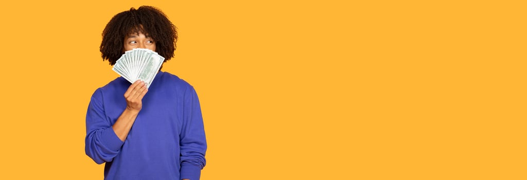 An man stands confidently, holding a fan of cash close to his face. They wear a casual blue sweater, showcasing a playful expression against a vibrant yellow backdrop, panorama, copy space photo