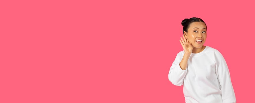 A smiling Asian woman wearing a white sweater waves enthusiastically, set against a vibrant pink backdrop, copy space photo