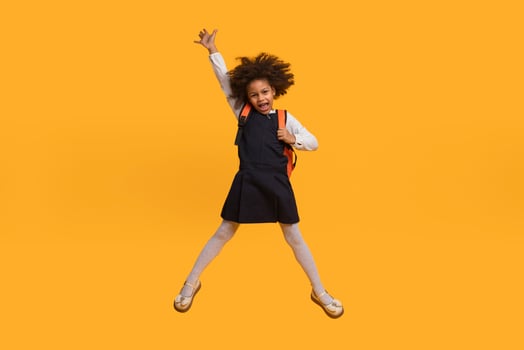 A young African American girl in a school uniform jumps in the air with her arms raised and a smile on her face. She is wearing a black dress, a white shirt, and a backpack photo