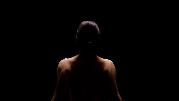 A woman sits with her back to the camera, illuminated by a single light source. She is facing away from the viewer and the background is completely black. photo