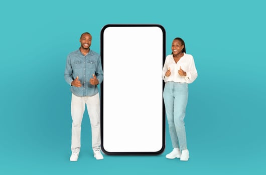 African American couple stand smiling and giving a thumbs-up next to an oversized smartphone with a blank white display photo