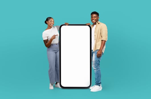 Black man and woman are standing against a blue background, holding up a blank screen. They appear to be showcasing something on the screen, potentially for a presentation or advertisement. photo