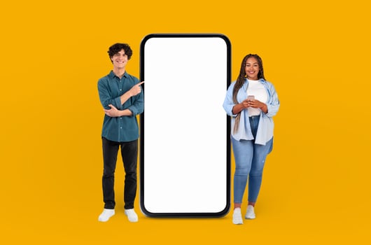 The vibrant yellow background highlights the smiling multiracial young couple as they gesture towards the expansive blank screen of the smartphone beside them. photo