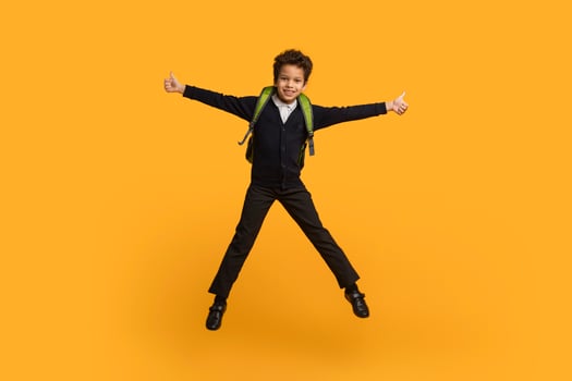 Black school boy, wearing a black uniform and a green backpack, jumps in the air with a big smile. He has his arms outstretched and is giving a thumbs up with both hands. photo
