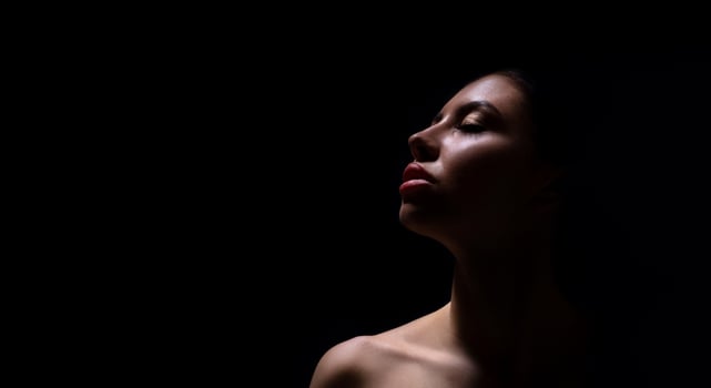A close-up photograph of a womans face, captured in a dark environment. The woman is facing upwards, and her face is illuminated by a single light source, casting shadows across her features. photo