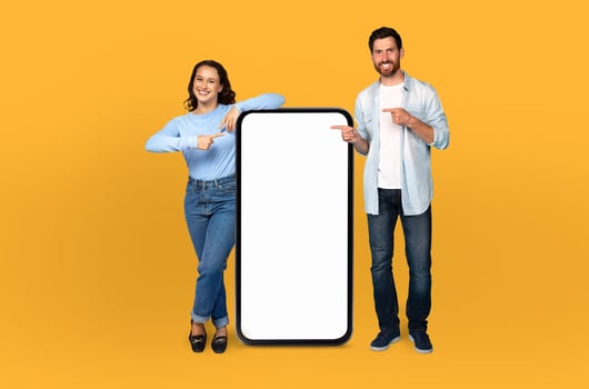 A man and woman are standing in front of a blank screen, both pointing towards it with their index fingers. The background is a solid yellow color, creating a vivid contrast with the neutral screen. photo