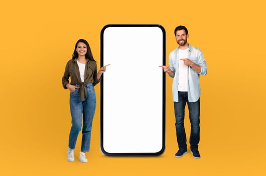 Multiethnic couple standing in front of a bright yellow background, both pointing at a blank screen. They seem focused and engaged photo