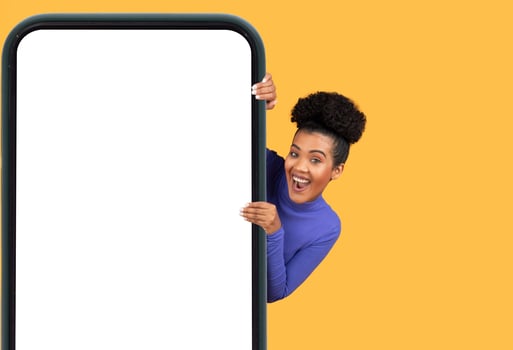 A young woman with curly hair joyfully leans out from behind a large mobile device, showcasing her excitement. The bright yellow backdrop adds a cheerful touch, enhancing the playful atmosphere photo