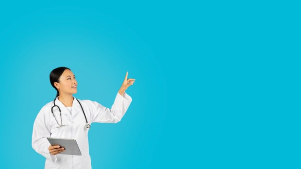 Asian woman doctor in a white coat, with a stethoscope around her neck, is smiling and holding a tablet in her left hand while pointing at copy space. She is in front of a solid blue background. photo