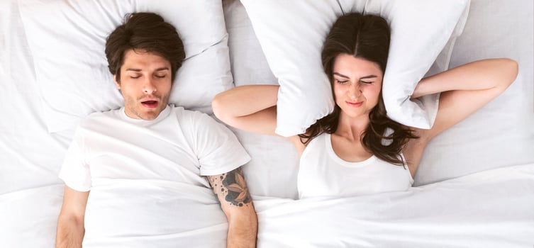 A woman lies in bed with her eyes closed and her hands covering her ears while a man sleeps beside her. The mans mouth is open as he snores loudly. The woman appears annoyed or frustrated. photo