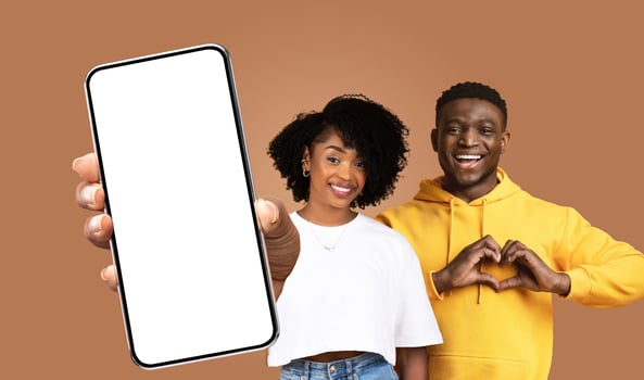 A young black woman and man stand against a brown background. The woman holds a smartphone with a blank screen. The man smiles and makes a heart shape with his hands. photo