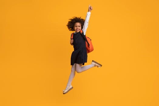 African American girl wearing a school uniform and backpack. She is jumping in the air with her arms raised, smiling, and giving a thumbs up. The background is a solid yellow color. photo