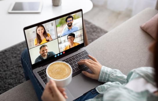 A woman sits on a couch with a laptop in her lap. She is participating in a call with three other people. The laptop screen shows the faces of the four participants in a grid format. photo