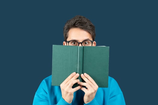 A young man sits against a solid dark blue background, holding an open book in front of his face. He wears glasses and a bright blue sweater, peering over the top of the book photo