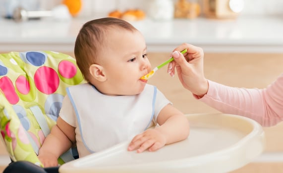 A woman is sitting and feeding a baby with a spoon. The baby is sitting on a high chair while eagerly eating the food. The woman gently guides the spoon into the baby mouth. photo