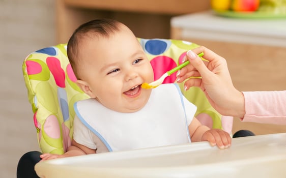 A happy baby sits in a colorful high chair while being fed with a spoon by an adult. The scene takes place in a bright home kitchen, suggesting a casual and joyful mealtime. photo