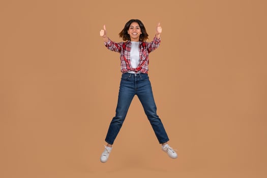 A young middle eastern woman with long, dark hair is jumping in the air with her arms outstretched and thumbs pointed upwards. She is wearing red and white plaid shirt, blue jeans, and white sneakers photo