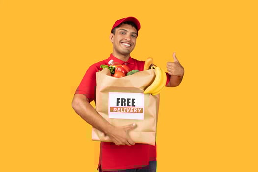 Delivery man wearing a red shirt is seen holding a bag of food, showing thumb up. He appears to be standing on yellow background. The bag is filled with groceries photo