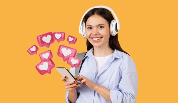 Smiling lady student in casual wear using her smartphone wearing white headphones, sending love message, carrying grey backpack, standing on yellow background photo