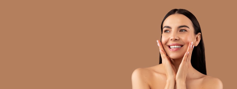 Aesthetic medicine and cosmetology concept. Happy beautiful brunette young woman touching her perfect face skin, posing naked over beige studio background, looking at copy space and smiling, panorama photo