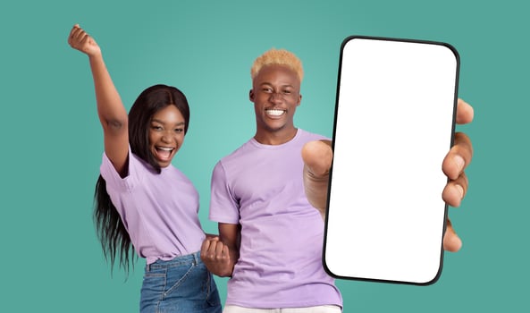 A young black couple in matching purple shirts stands in front of a teal background. The man holds a smartphone with a blank white screen towards the camera, while the woman celebrates success photo