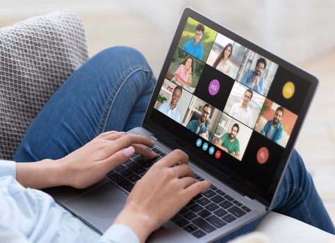 Lady sitting on a couch, participating in a virtual business meeting with multiple colleagues using a laptop. The screen shows various participants engaged in the discussion on a conference photo