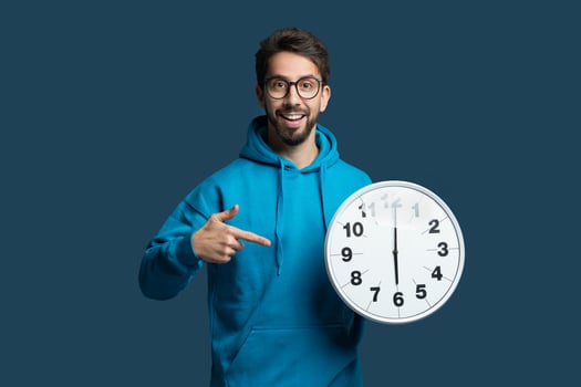 A young man in a blue hoodie stands against a blue background, holding a round wall clock. He points excitedly at the clock, showcasing the time while expressing a joyful and enthusiastic demeanor. photo