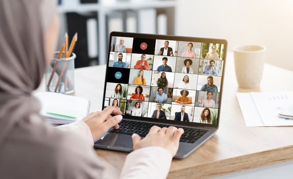 Muslim woman sits at a desk in a home office setting, participating in a large virtual meeting on her laptop. The screen is filled with tiles of diverse people participating in the meeting photo