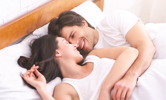 A couple lies in bed, a man and a woman, both smiling and looking at each other. The woman lies on her back with her head resting on the pillow, while the man is on his side, looking down at her photo
