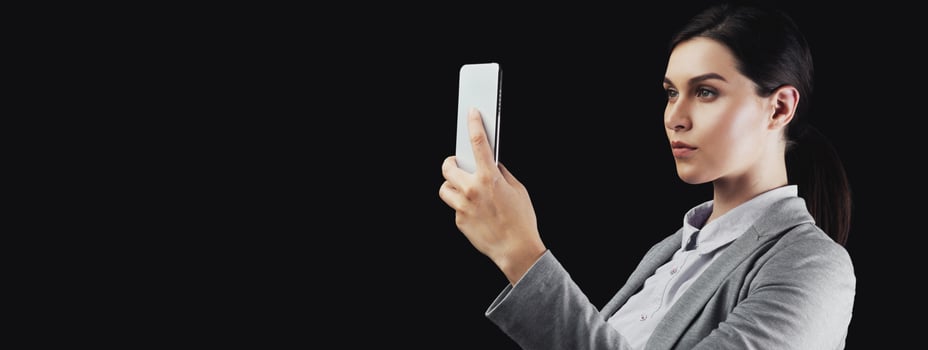 A woman in a grey blazer takes a selfie with her smartphone against a black background. photo