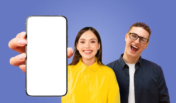 A woman holds a smartphone with a white blank screen in front of her, smiling at the camera. A man wearing glasses and a dark shirt stands behind her, smiling and looking at the camera. photo