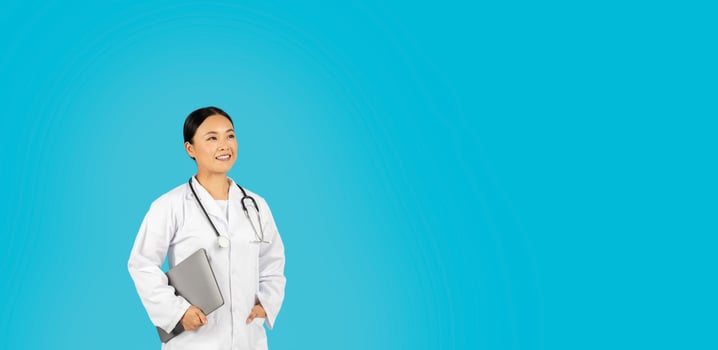 Asian woman doctor, wearing a white coat and a stethoscope, smiles while holding a laptop in her left hand and looking at copy space. She is standing in front of a light blue background. photo