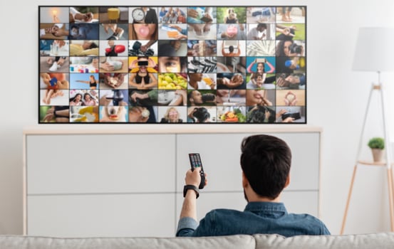 A man sits on a couch and watches television, holding the remote in his right hand. The TV screen displays a collage of various images, including people, objects, and scenes photo