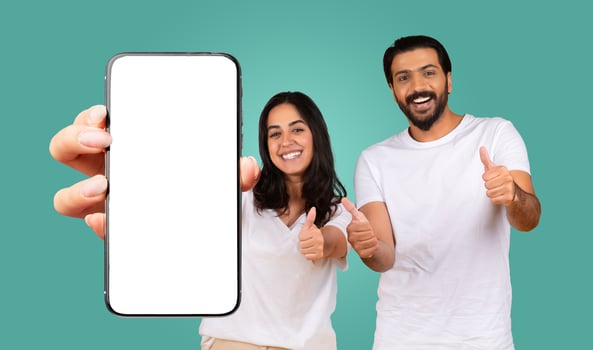 A happy Eastern young couple with dark hair, wearing white t-shirts, gives a thumbs-up while holding a smartphone with a white screen. The couple is standing in front of a turquoise background. photo