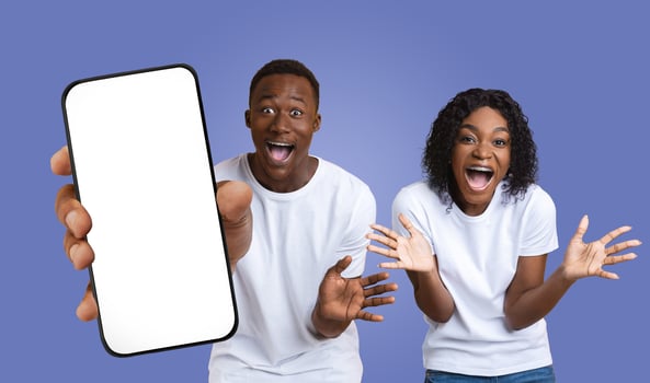 A young black couple, dressed in casual white shirts, express their excitement while holding a smartphone with a blank screen. The background is a solid purple color. photo