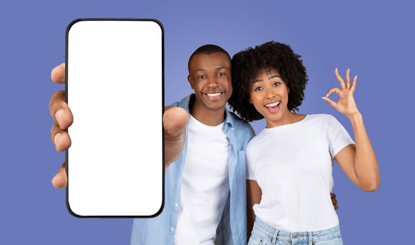 A young African American couple stands together in front of a purple background, smiling and looking at the camera. The man is holding a smartphone with a blank screen, woman showing okay photo