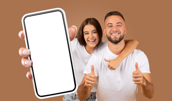 A happy young couple stands in front of a brown background, holding a smartphone with a blank screen and giving thumbs up, showcasing their excitement and approval. photo