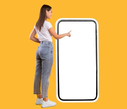 Full Body Length Back View Of Woman Using Big Smartphone With Blank White Screen And Touching Display Panel With Finger, Cheerful Lady Standing On Yellow Background, Ordering Food Delivery, Mock Up photo