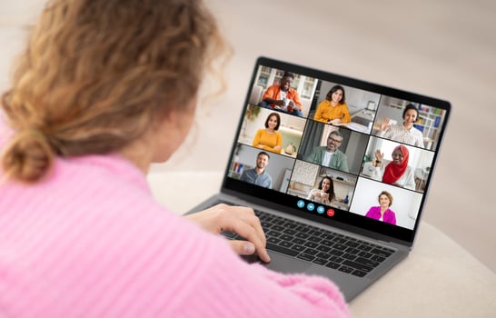 A woman is sitting at a desk and looking at a laptop screen. She is participating in a conference call with eight other people. The people on the screen are in a grid layout photo