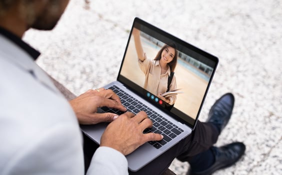 African American man sits outdoors, using his laptop to chat with a woman. He is typing on the keyboard and smiling, while the woman on the screen waves and holds books. photo