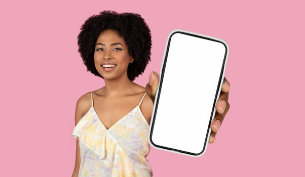 African American woman smiles brightly at the camera while holding a smartphone with a blank screen in front of a pink background. photo