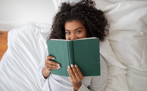 Hispanic woman is laying comfortably in bed, engrossed in a book she is reading. The room is softly lit, top view photo